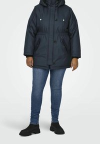Marinblå vadderad parkas med huva, frambutcher, två sidofickor och elastisk midja. Bärs över blå jeans och svarta ankelstövlar.