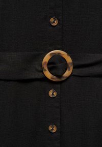 Camicia in tessuto nero con una grande fibbia in tartaruga. Sei bottoni di diverse tonalità di marrone sono allineati verticalmente sul davanti.