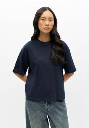 Jeune femme aux longs cheveux foncés portant un T-shirt ample bleu marine et un jean bleu clair, debout les mains dans le dos.
