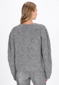 Szürke kötött pulóver, textúrált dizájnnal, hosszú ujjakkal és laza szabással. Az anyag lágy és enyhén bolyhosnak tűnik.