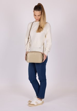 Žena s dlouhými vlasy v bílém svetru, modrých džínách a bílých teniskách drží oběma rukama před sebou béžovou kabelku přes rameno.