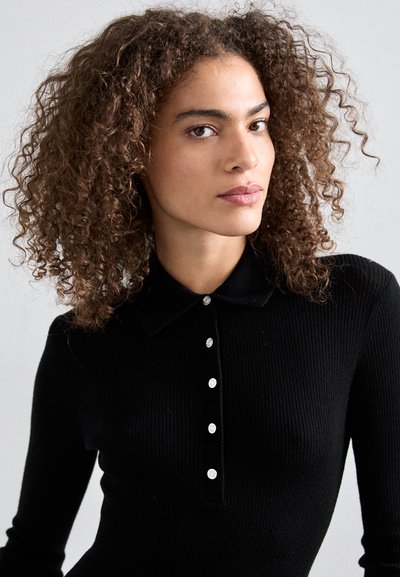 Polo shirt noir à côtelage avec un col et cinq boutons métalliques sur le devant. Le tissu semble ajusté et texturé avec une finition lisse.