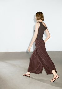 Top asimmetrico marrone abbinato a pantaloni larghi in satin marrone e sandali neri minimalisti, su uno sfondo neutro.