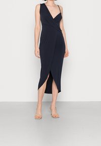 Kobieta w granatowej sukience midi z asymetrycznym dekoltem i rozcięciem z przodu, w połączeniu z beżowymi sandałami na obcasie, stojąca przed białą ścianą.