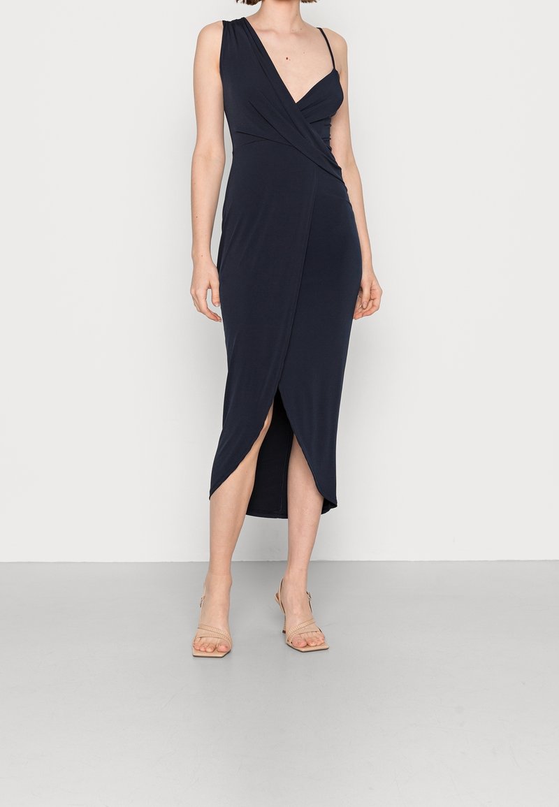 Kobieta w granatowej sukience midi z asymetrycznym dekoltem i rozcięciem z przodu, w połączeniu z beżowymi sandałami na obcasie, stojąca przed białą ścianą.