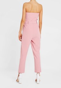 Combinaison rose sans bretelles avec un corsage ajusté, une taille ceinturée et des jambes fuselées. Le matériau semble lisse avec une finition mate. Sandales à talons blocs blanches.