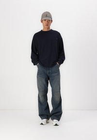 Ναυτικό μπλουζάκι μακρύ μανίκι με τσέπη στο στήθος, συνδυασμένο με ξεθωριασμένα μπλε τζιν που έχουν λεπτομέρειες από μπαλώματα, και λευκά sneakers με πράσινες πινελιές.