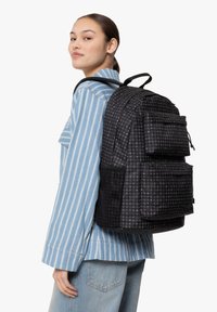 Mochila negra con patrón a cuadros, múltiples bolsillos frontales y correas ajustables. Llevada sobre el hombro, combinada con una camisa a rayas.