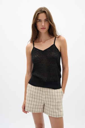 Žena v čiernom priehľadnom tielku bez rukávov a béžových kockovaných šortkách stojí s jednou rukou v vrecku na bielom pozadí.