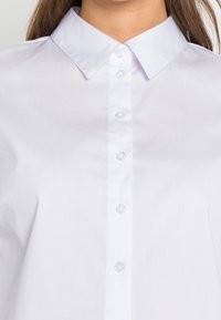 Hvid skjorte med knaplukning og en skarp krave, med en række af klare knapper, glat tekstur og en struktureret design.