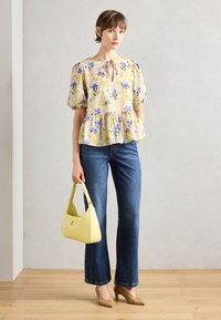 Blumen-Peplum-Bluse in Gelb mit lila Akzenten, mit Puffärmeln und Keyhole-Ausschnitt. Kombiniert mit ausgestellten Jeans und einer gelben Handtasche.