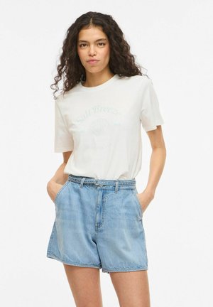 Femme aux cheveux bouclés portant un t-shirt blanc à motif et un short en jean bleu clair taille haute, les mains dans les poches.