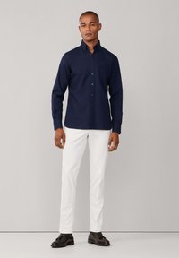 Camicia blu navy con bottoni e maniche lunghe, abbinata a pantaloni bianchi slim fit e scarpe marrone scuro, creando un look pulito e moderno.
