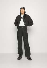 Black bomber jacket over a white shirt, paired with black pinstripe pants featuring side stripes and a black cap, standing with hands on hips.