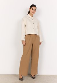 Veste beige cintrée avec des boutons argentés, associée à un pantalon large beige et décontracté. Des escarpins à rayures noir et blanc complètent l'ensemble.