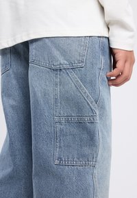 Light blue denim pants with a relaxed fit, featuring large side pockets and a textured finish. The fabric shows subtle variations in color.