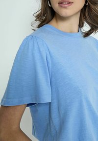 Šviesiai mėlyna trumpa rankovė t-shirt su lygiu paviršiumi ir laisvu siluetu. Su pūstomis rankovėmis ir apvaliu iškirpimu, skirtas kasdieniam įvaizdžiui.