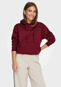 Felpa con cappuccio bordeaux con tasca frontale, polsini a coste e vestibilità comoda, abbinata a pantaloni beige con strisce laterali.
