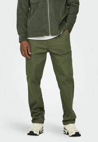 Pantalones cargo verde oliva hechos de una tela texturizada, con un corte ajustado y bolsillos laterales, combinados con zapatillas de color claro.