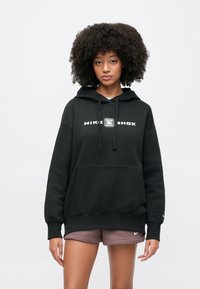 Fiatal nő göndör hajjal, fekete Nike Shox kapucnis pulóvert és mályva Nike rövidnadrágot viselve, egyszínű fehér háttér előtt áll.