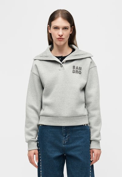 Γυναίκα που φοράει ανοιχτό γκρι φούτερ με γιακά και το κείμενο "SANDRO" στο στήθος, συνδυασμένο με μπλε τζιν με τρούκς στο πλάι.