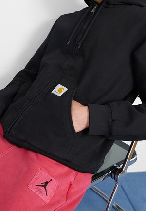 Person trägt schwarzen Kapuzenpullover mit Reißverschluss und Carhartt-Logo sowie pinke Hose mit schwarzem Jordan-Logo und sitzt auf einem verchromten Stuhl.