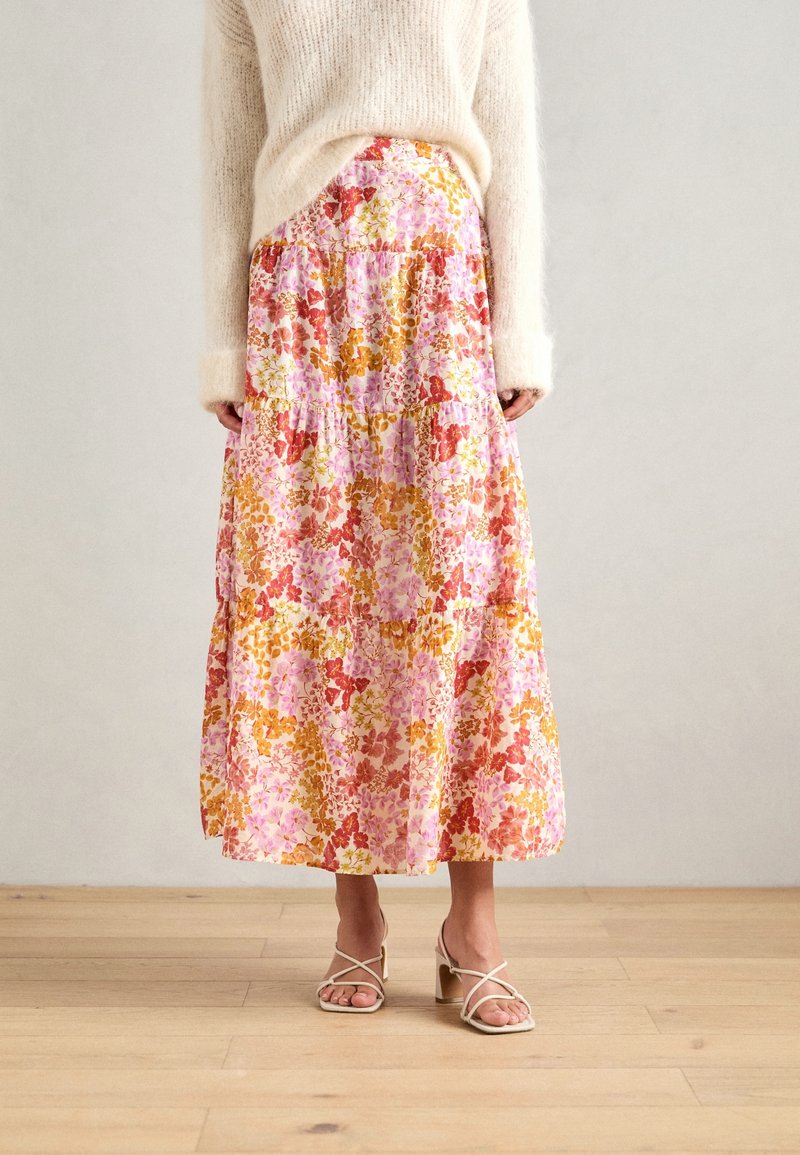 Jupe maxi à motifs floraux dans des teintes de rose et d'orange, avec un design à volants et un tissu léger. Associée à des sandales beiges à lanières.