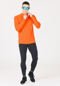 Klarr orange långärmad träningsskjorta, svarta åtsittande tights och svarta löparskor. Blå sportglasögon accentuerar outfiten.