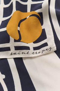 Zīda šalle ar tīkla dizainu tumši zilā un krēma krāsā, ar drosmīgu dzeltenu abstraktu formu. Iekļauts izšūts etiķetes uzraksts "saint tropez".