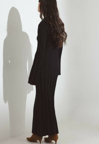 Ženska z dolgimi valovitimi lasmi, oblečena v črno rebrasto pleteno obleko in rjavimi gležnjarji, stoji bočno in meče senco na belo steno.