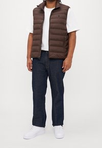 Brun pufferväst med hög krage över en vit t-shirt, kombinerad med mörkblå jeans och vita sneakers. Enkel design, inga synliga mönster.