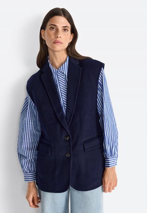 Navy blue wool-blend vest with peaked lapels, single button closure, and two front pockets, layered over a blue and white striped shirt.