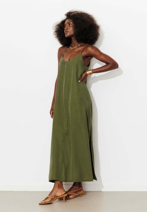 Femme avec coiffure afro, robe longue sans manches vert olive, collier et bracelets en or, portant des sandales beige à talons bas et brides.