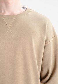 Světle krémový svetr s kulatým výstřihem, volným střihem a viditelnými stehy přes rameno. Měkká textura.