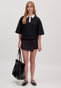 Zwarte oversized polo shirt met een witte kraag, gecombineerd met een zwarte mini rok, terwijl ze een glanzende zwarte tote bag vasthoudt en zwarte loafers draagt.
