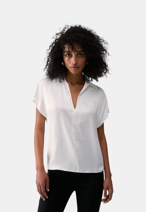 Donna con capelli ricci indossa una camicia bianca a maniche corte con colletto e scollo a V profondo, abbinata a pantaloni neri, su sfondo semplice.