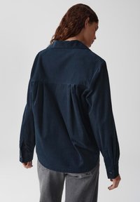 Donna con capelli ricci che indossa una maglietta con maniche lunghe di colore blu scuro e pantaloni grigi, vista di spalle su uno sfondo chiaro e semplice.
