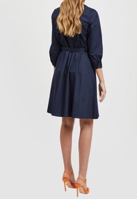 Mörkblå, figursydd klänning med ärmar som går till armbågen och midja med bälte. Klänningen har en utsvängd kjoldesign och bärs med orange högklackade sandaler.