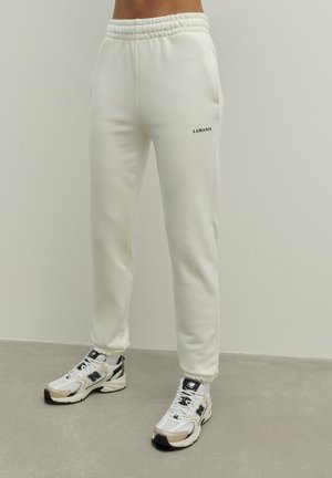 Osoba ubrana w białe spodnie dresowe LAMANIA z elastycznymi ściągaczami przy kostkach i w pasie, w zestawieniu z biało-beżowymi butami sportowymi New Balance, stojąca na szarej podłodze.