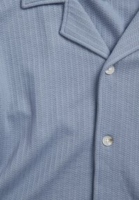 Lichtblauw kortmouwshirt van een gestructureerde, ribbelachtige stof. Voorzien van een notch-kraag en twee witte knopen langs de knoopsluiting.