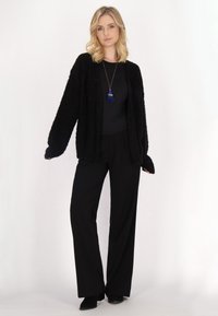 Cardigan negru, pufoasă, peste un top negru, asortată cu pantaloni largi negri. Mâneci lungi și o croială lejeră. Accentuata cu un colier cu ciucure albastru.