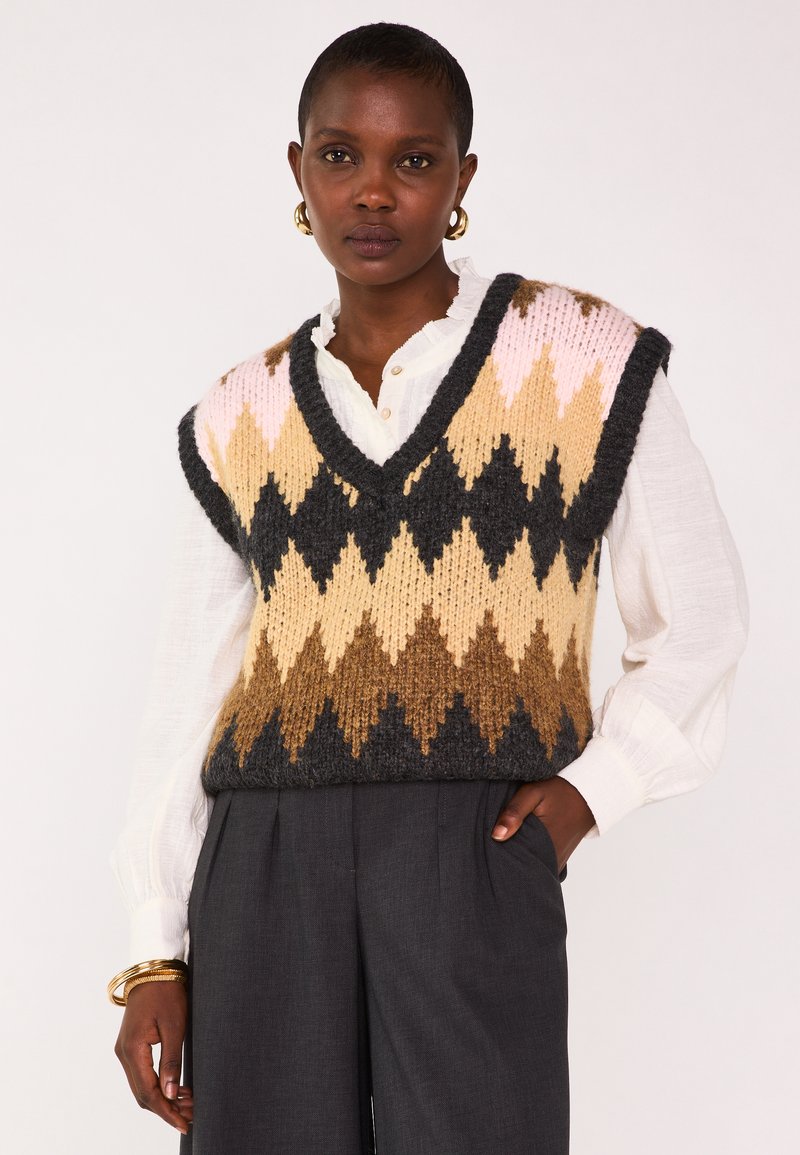 Pull sans manches en tricot dans un motif en zigzag rose, beige, marron et noir ; porté par-dessus une chemise blanche à col avec des détails de boutons.