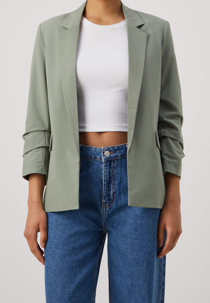 Blazer verde chiaro, su misura, con colletto a punta e maniche arrotolate, abbinato a un top bianco corto e jeans blu a vita alta.