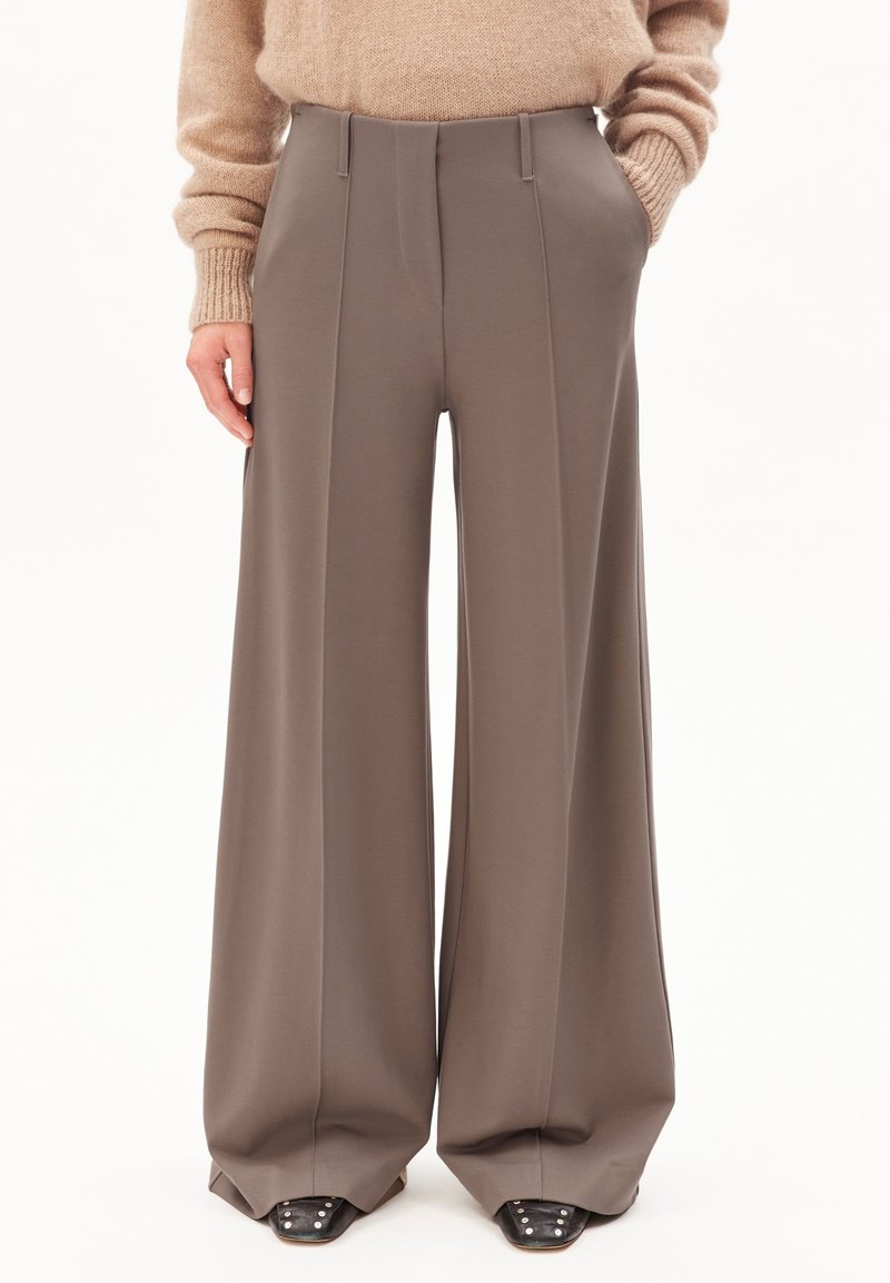 Personne portant un pantalon taupe à jambes larges et un pull en maille beige, les mains partiellement dans les poches et des chaussures noires cloutées visibles.