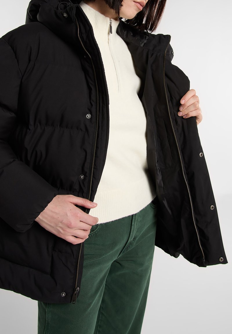 Chaqueta acolchada negra con capucha, con cierre de cremallera y botones a presión. Llevada sobre un suéter blanco de cuello alto y pantalones de pana verdes.