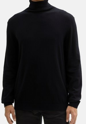 Homme portant un pull à col roulé noir uni à manches longues, debout devant un fond blanc.