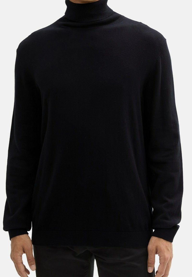 Homme portant un pull à col roulé noir uni à manches longues, debout devant un fond blanc.