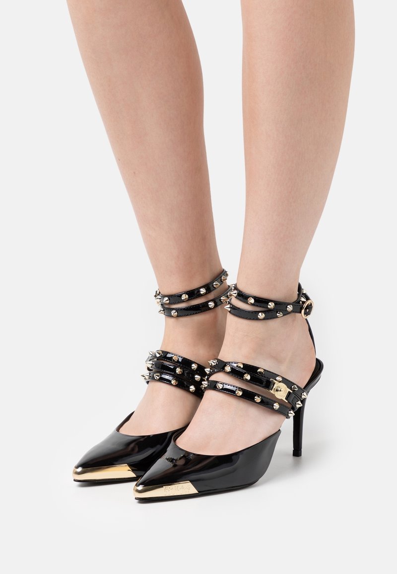 Escarpins pointus en cuir verni noir avec embouts métalliques dorés et brides à clous autour de la cheville et du pied, portés sur des jambes nues.