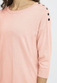 Pull en tricot rose avec un col rond et des manches trois-quarts. Comprend des boutons décoratifs noirs le long de la couture de l'épaule. Texture douce.