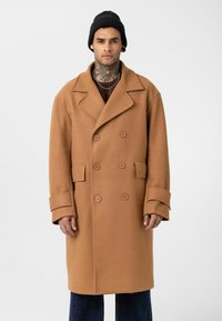 Man met een nektatoeage, gekleed in een zwarte muts, een camel kleurige double-breasted jas en donkere broek, staand tegen een effen witte achtergrond.